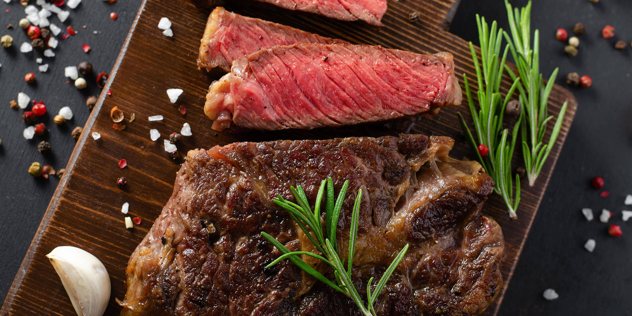 Hand-cut ribeye steak on a butcher board with rosemary and peppercorns